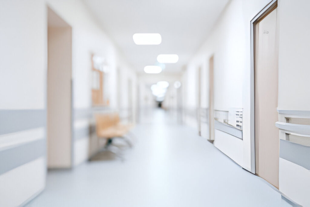 A bright, clean hospital hallway with white walls, light floors, and closed doors on both sides. Empty chairs line the wall—a calm scene where patients may await evaluation for Dupuytren’s contracture symptoms. Overhead lights softly illuminate the corridor.