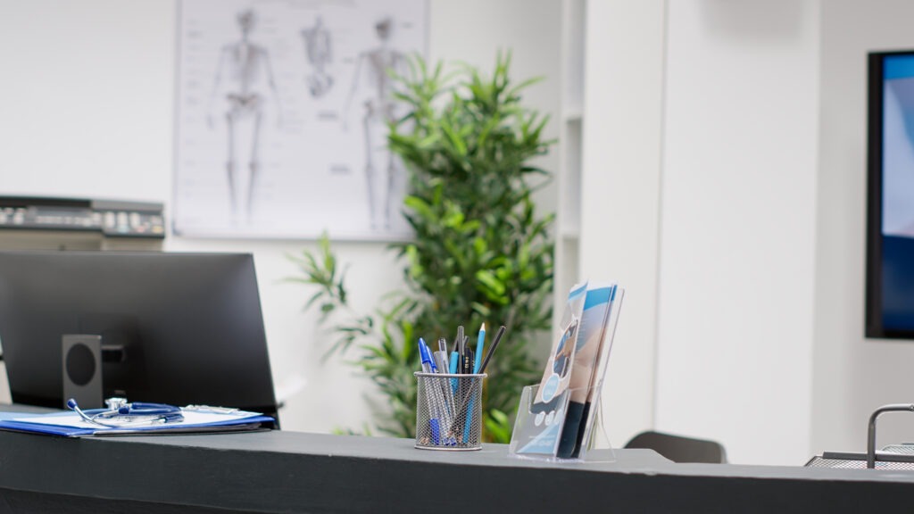 A modern medical office reception desk with a computer, clipboard, pens in a holder, and brochures about Dupuytren’s contracture symptoms. A potted plant and anatomical posters are visible in the background.