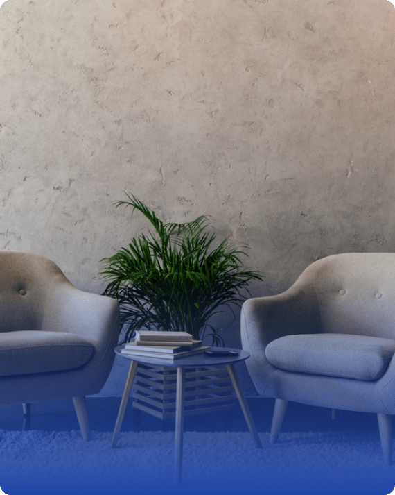 Two light gray armchairs face each other with a small round wooden table in between, holding stacked books about Dupuytrens disease. A green potted plant sits behind the table, against a textured beige wall.