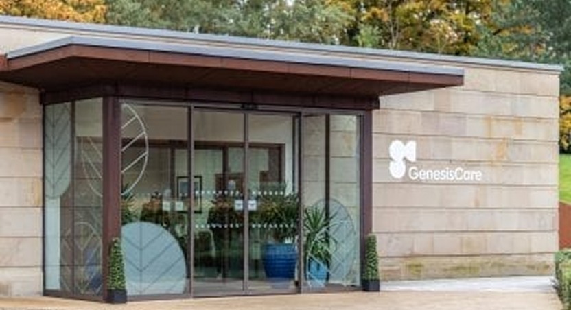 The entrance of a GenesisCare medical facility with glass doors, large windows, and a modern design, where patients can seek expert care for conditions like Dupuytren’s contracture symptoms. The GenesisCare logo is displayed on the beige stone wall near the entrance.