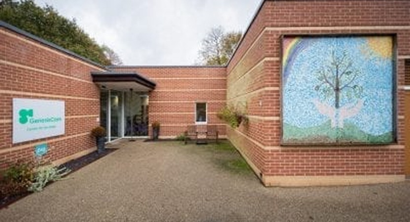 A brick building with a glass entrance and a large mosaic mural on the right wall, depicting a tree, a white bird, and a rainbow. A sign near the entrance displays the GenverClem logo—experts in Dupuytrens contracture treatment UK. Shrubs line the walkway.