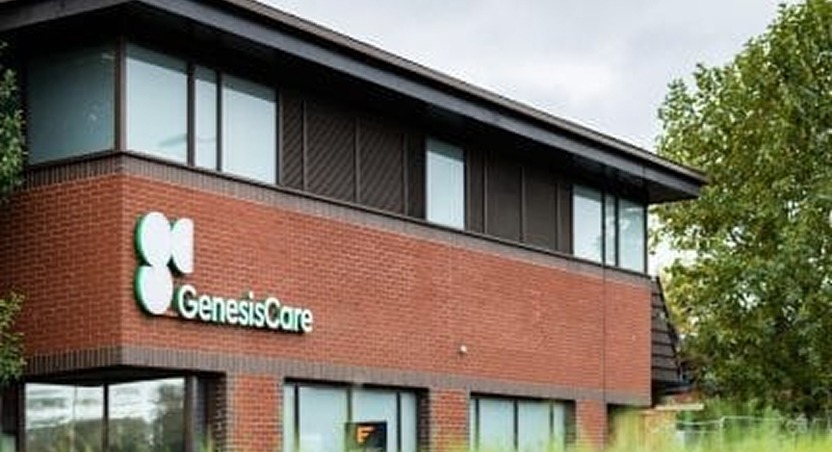A two-story brick building with large windows and a GenesisCare sign on the exterior wall, surrounded by trees and greenery. The clinic offers dupuytrens contracture treatment UK in a modern and welcoming setting.