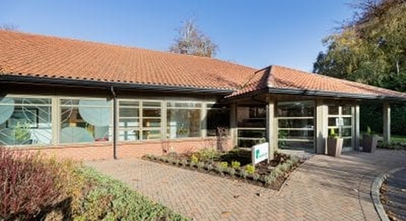 A modern single-story building with large windows, a red-tiled roof, and landscaped garden beds at the entrance. The clear blue sky mirrors the tranquility often sought by those researching Dupuytren’s contracture symptoms. Trees are visible in the background.