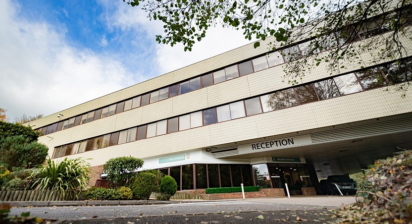 A three-story building with large windows and a sign for Reception above the entrance, surrounded by greenery and trees—much like how Dupuytrens contracture symptoms can gradually surround and affect the hand—viewed from a low angle under a partly cloudy sky.