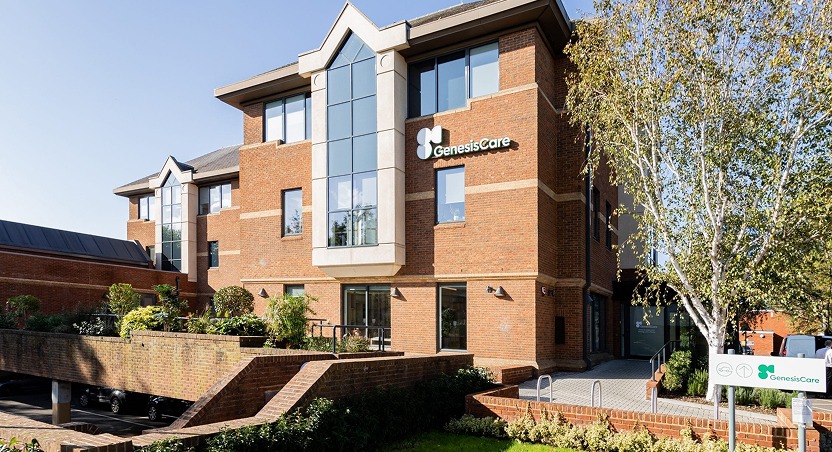 A modern brick building with large glass windows and a GenesisCare sign, offering Dupuytren’s contracture treatment UK, surrounded by greenery and trees under a clear blue sky.