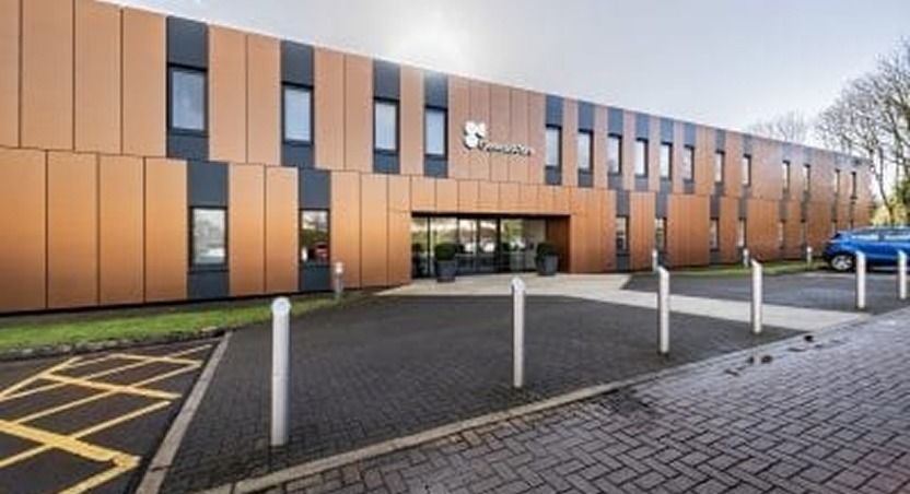 A modern two-story building with brown and black panels, large windows, and a main entrance. Near the sidewalk and parking spaces, where a blue car is parked, banners about Dupuytrens disease awareness are visible among the trees in the background.
