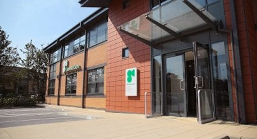 Modern brick office building with large windows and glass entrance; a sign with a green logo is displayed beside the door. The facility provides expert Dupuytrens contracture treatment UK. Empty parking spaces and trees are visible outside.
