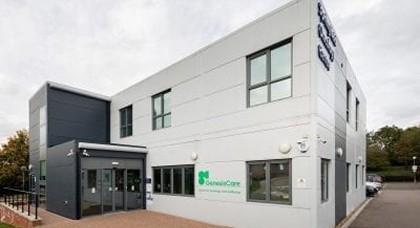 A modern, two-story building with large windows and a gray exterior. Signs near the entrance and on the wall read GenesisCare, a center specializing in conditions like dupuytrens contracture disease. Trees and a cloudy sky are in the background.