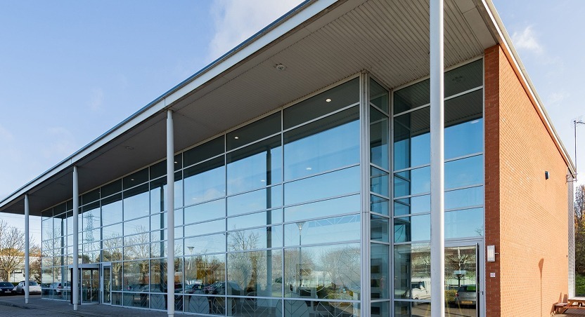 A modern building with large glass windows and a flat roof, featuring a red brick wall on one side. The sunlight reflects off the windows, and there are cars and trees outside—much like clinics offering Dupuytrens contracture treatment UK.