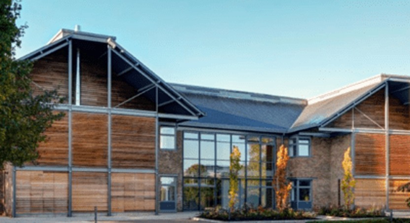 Modern building with large glass windows, wood and brick exterior walls, and a sloped metal roof, surrounded by small trees—much like innovative spaces for dupuytrens contracture treatment UK—set under a clear blue sky.