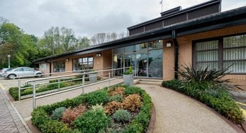 Single-story brick building with large windows and a glass entrance, ideal for those seeking Dupuytrens contracture treatment UK. A curved path with a handrail leads to the door, bordered by neat shrubs. Parked cars and trees are seen in the background.