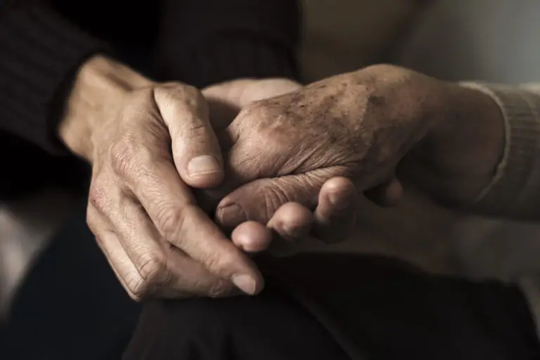 A younger person gently holding the hand of an older person, showing support and care. The image focuses on their hands, highlighting the contrast in age, warmth of human connection, and visible signs of Dupuytren’s contracture symptoms.