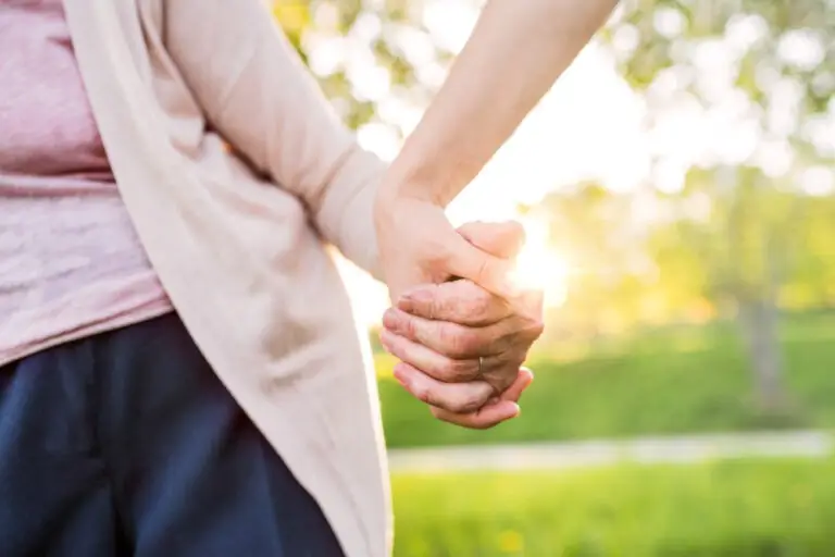 Close-up of two people holding hands outdoors with sunlight shining through, suggesting warmth and connection. The blurred green background evokes a park setting—an uplifting scene that resonates with those seeking Dupuytrens contracture treatment UK.