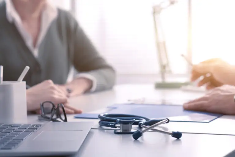 Two people sit across from each other at a desk in an office. A stethoscope, glasses, and clipboard rest on the desk, suggesting a medical consultation about Dupuytren’s contracture symptoms or causes of Dupuytren’s disease is taking place.