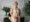 A woman with short blonde hair, wearing a beige blazer, stands indoors holding eyeglasses. She smiles softly, with a side table and green leafy plants in the background—her calm demeanor belies any concerns about Dupuytren’s contracture symptoms.