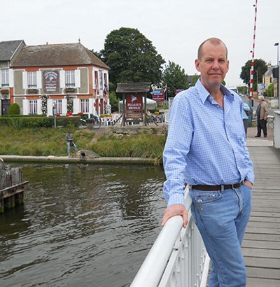 A man in a blue checkered shirt and jeans, affected by Dupuytrens contracture disease, leans on a white railing of a bridge over a river, with buildings, trees, and a sign visible in the background on a cloudy day.