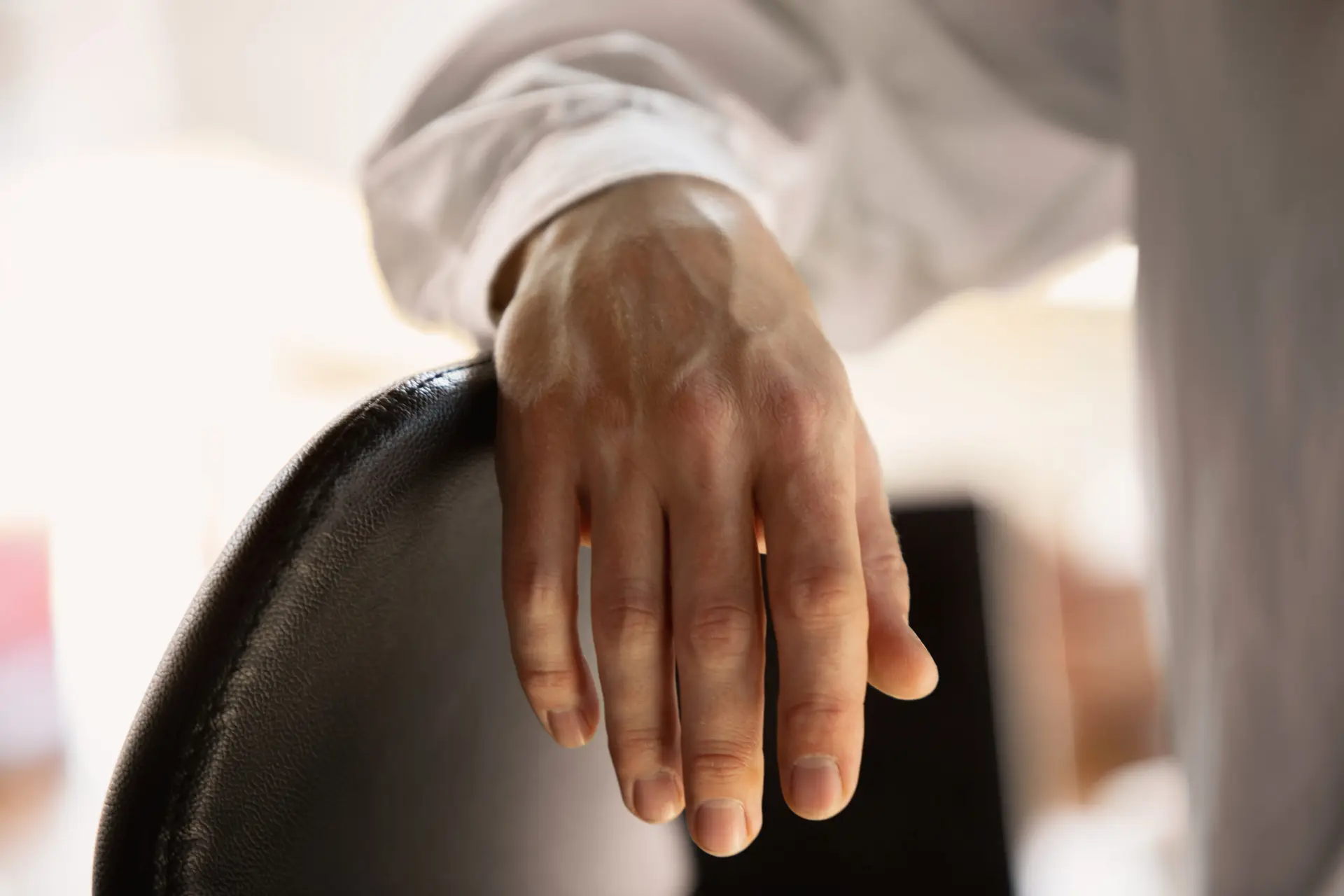 A close-up of a person’s hand, possibly affected by Dupuytren’s contracture symptoms, resting on the back of a black chair. The person is wearing a white long-sleeve shirt and the background is softly blurred.