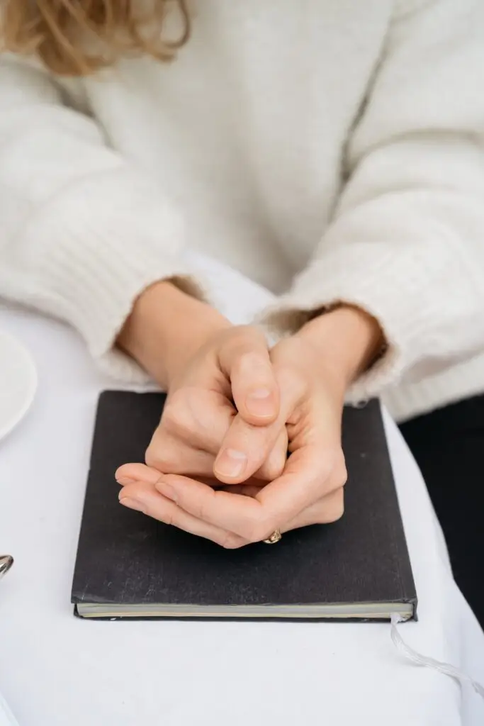 A person in a white sweater sits at a table with hands clasped together, possibly affected by Dupuytrens contracture disease, resting on a closed black notebook.