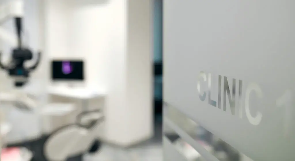 A blurry view of a modern medical clinic room with a computer, exam chair, and microscope hints at advanced care like Dupuytrens contracture treatment UK. The word CLINIC is partially visible on a frosted glass door in the foreground.