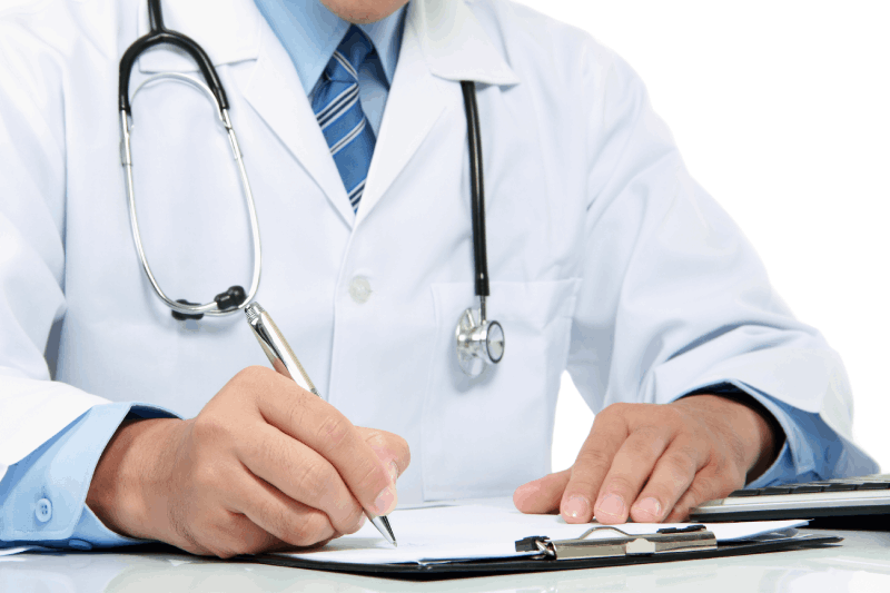 A doctor in a white coat with a stethoscope around their neck writes on a clipboard at a desk, possibly reviewing causes of Dupuytrens disease, with only their torso and hands visible.