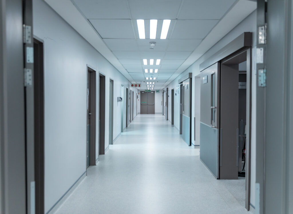 A clean, empty hospital corridor with closed doors on both sides hints at specialized care areas, possibly for conditions like Dupuytren’s contracture symptoms or Ledderhose disease. The bright lights and shiny floor enhance the sterile, quiet atmosphere.