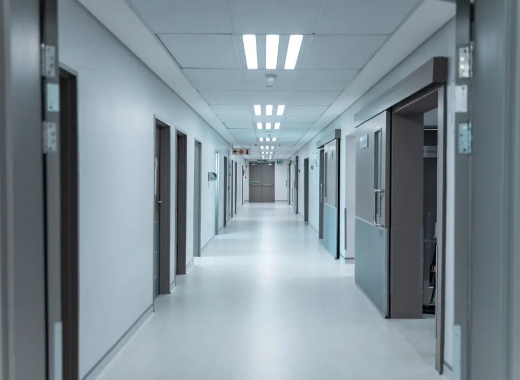 A clean, empty hospital corridor with closed doors on both sides hints at specialized care areas, possibly for conditions like Dupuytren’s contracture symptoms or Ledderhose disease. The bright lights and shiny floor enhance the sterile, quiet atmosphere.