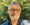 A man with short gray hair, a beard, and round glasses smiles outdoors. He is wearing a dark shirt, enjoying the sunlight filtering through green trees after his successful Dupuytrens contracture treatment UK.