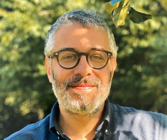 A man with short gray hair, a beard, and round glasses smiles outdoors. He is wearing a dark shirt, enjoying the sunlight filtering through green trees after his successful Dupuytrens contracture treatment UK.