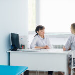A doctor and a patient are sitting across from each other at a desk in a bright, modern medical office, discussing Dupuytren’s contracture symptoms. The doctor is facing the patient, who is seated on a white chair.