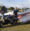 A person stands outdoors on grass, smiling beside a silver airplane and a black motorcycle, with a green shed and trees in the background under a partly cloudy sky, possibly reflecting on their experience with Dupuytren’s contracture symptoms.