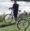 A person wearing a helmet and sunglasses stands with a bicycle near a grassy shoreline, waves behind them. Enjoying the sunny, windy day, they know staying active is important while managing Dupuytrens contracture disease. Another bicycle rests nearby.