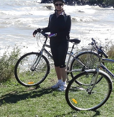 A person wearing a helmet and sunglasses stands with a bicycle near a grassy shoreline, waves behind them. Enjoying the sunny, windy day, they know staying active is important while managing Dupuytrens contracture disease. Another bicycle rests nearby.