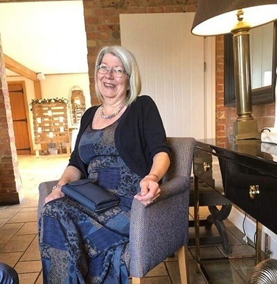 An older woman with gray hair, glasses, and a cheerful smile sits on a chair indoors, wearing a patterned blue dress and holding a navy clutch. She shows no visible Dupuytren’s contracture symptoms as she relaxes in the cozy brick-walled room.