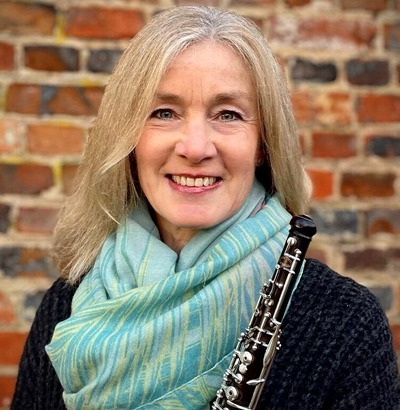 A smiling woman with long gray hair, wearing a light blue scarf and dark sweater, stands in front of a brick wall holding a clarinet—her joy undimmed despite living with Dupuytren’s contracture symptoms.