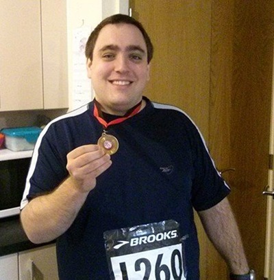 A smiling man wearing a dark sports shirt and a race bib holds up a medal in a kitchen, celebrating his achievement despite recently managing Dupuytren’s contracture symptoms.