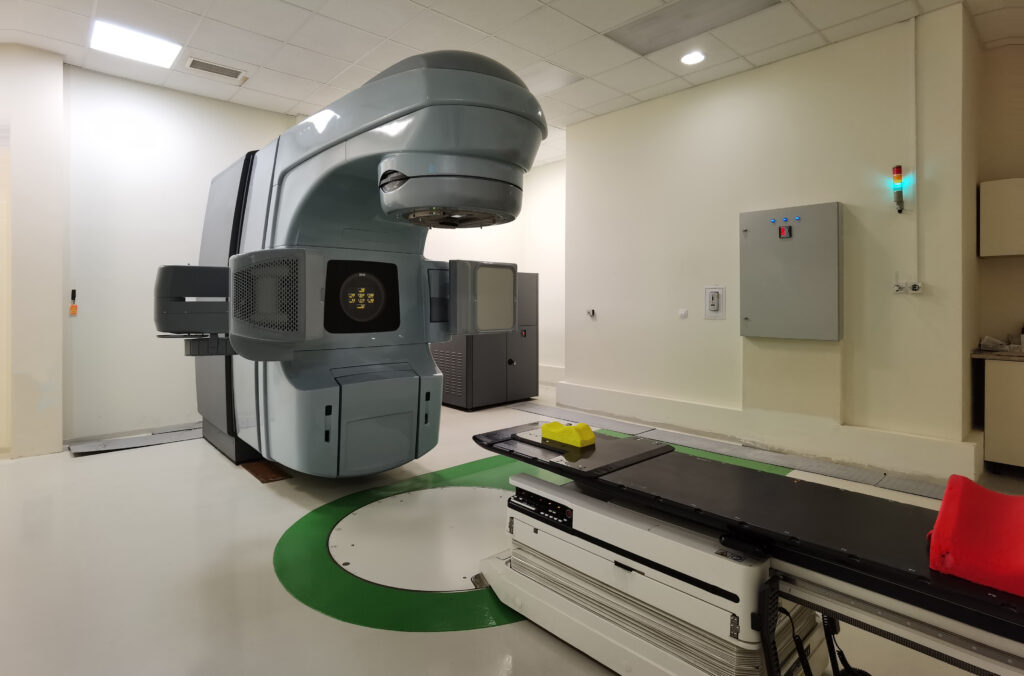 A large medical linear accelerator sits in a clean, well-lit radiation therapy room, used for treating conditions like Dupuytren’s contracture symptoms, with a treatment couch under the machine and safety indicators on the walls.