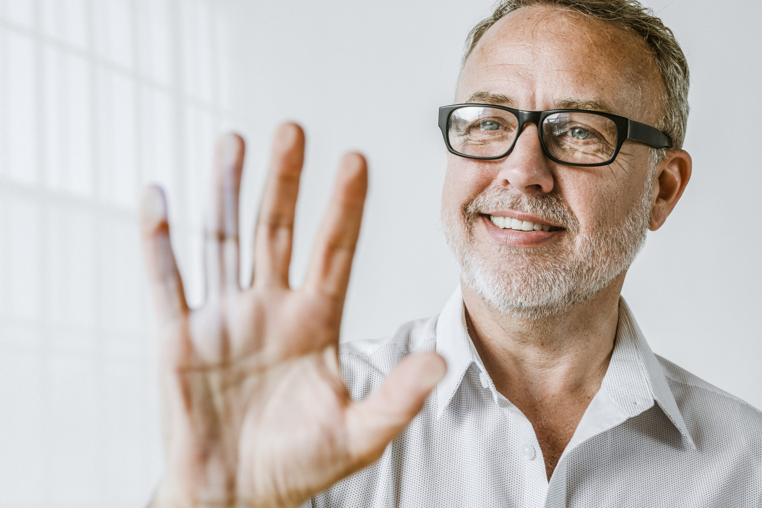 A smiling middle-aged man with glasses and a gray beard holds his hand up toward the camera, showing no visible Dupuytren’s contracture symptoms. He is wearing a light-colored shirt and standing in a bright, white room.