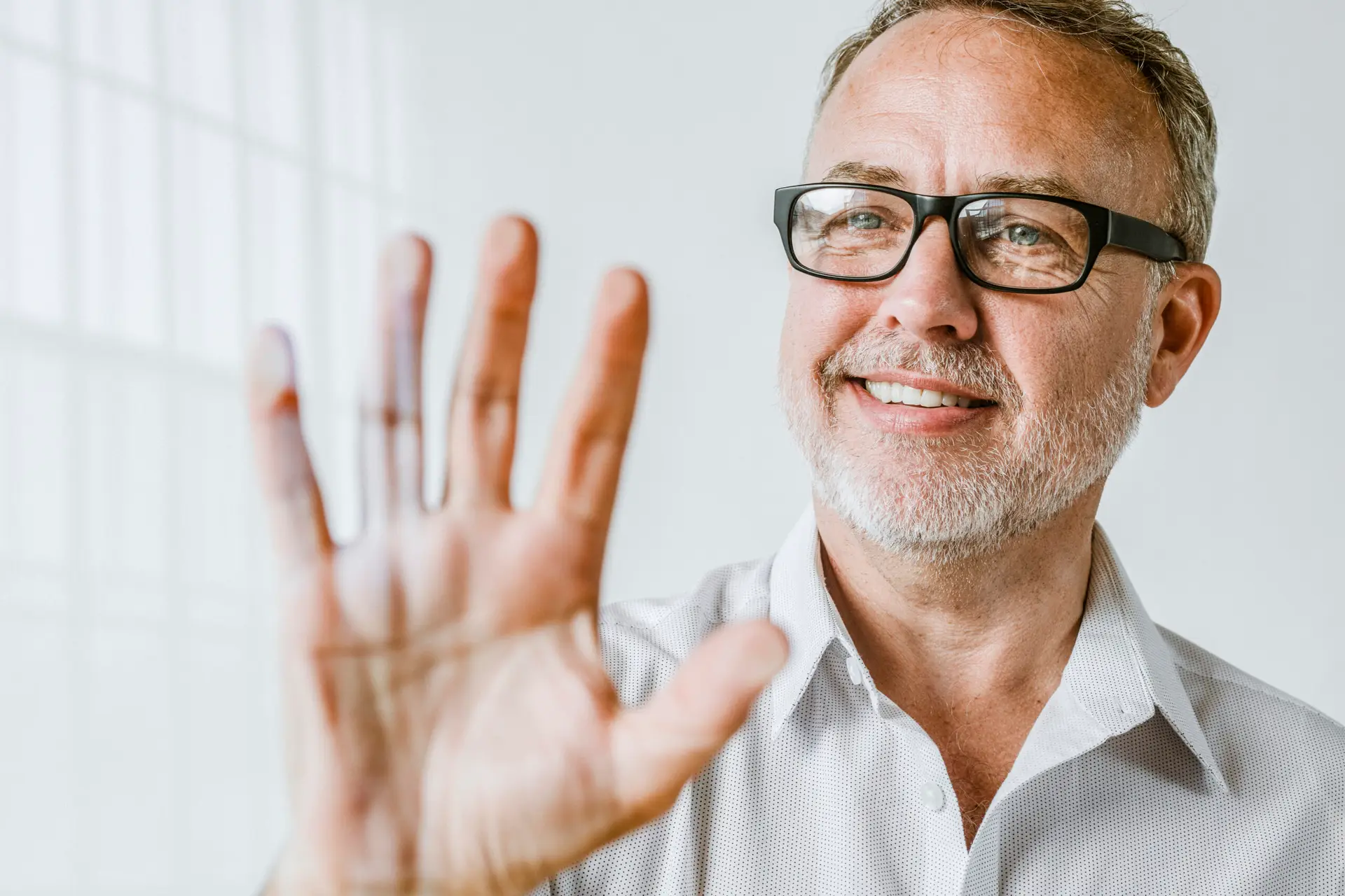 A smiling middle-aged man with glasses and a gray beard holds his hand up toward the camera, showing no visible Dupuytren’s contracture symptoms. He is wearing a light-colored shirt and standing in a bright, white room.
