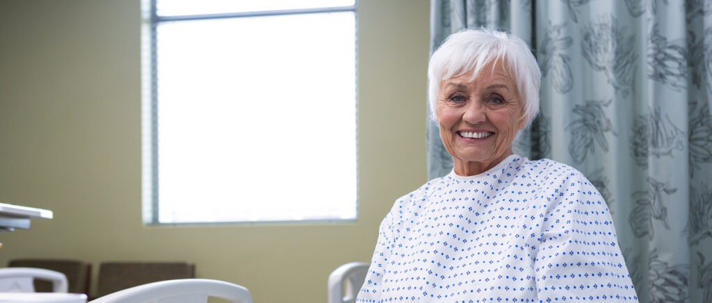 An older woman with short white hair, wearing a hospital gown, sits and smiles in a bright hospital room with a window and floral curtains in the background, perhaps awaiting treatment for Dupuytren’s contracture symptoms.
