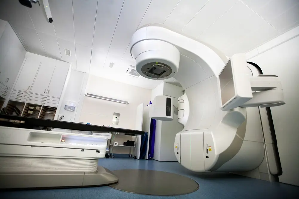 A modern hospital room features a large white medical machine for radiation therapy, often used in Dupuytrens contracture treatment UK, with an adjustable treatment table and organized cabinets along the wall. The space is well-lit and orderly.