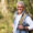 A smiling middle-aged man with short gray hair, wearing a white shirt, light gray vest, watch, and backpack, stands outdoors in a sunlit, wooded area—unaware of potential Dupuytren’s contracture symptoms that can affect hand mobility.