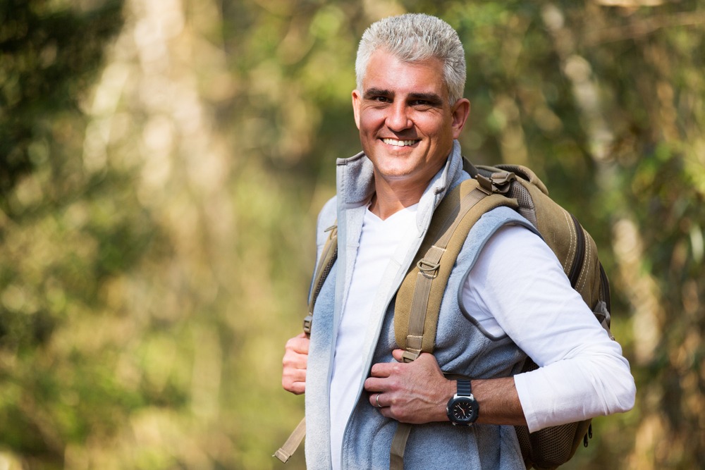 A smiling middle-aged man with short gray hair, wearing a white shirt, light gray vest, watch, and backpack, stands outdoors in a sunlit, wooded area—unaware of potential Dupuytren’s contracture symptoms that can affect hand mobility.