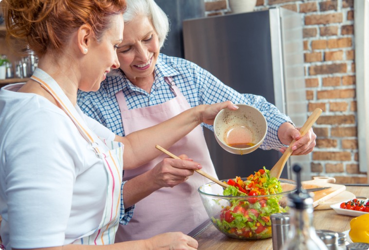 Two women in aprons prepare a fresh salad together, mixing colorful vegetables as one pours dressing and the other stirs. They’re chatting about healthy habits and exploring the causes of Dupuytrens disease in their daily routines.