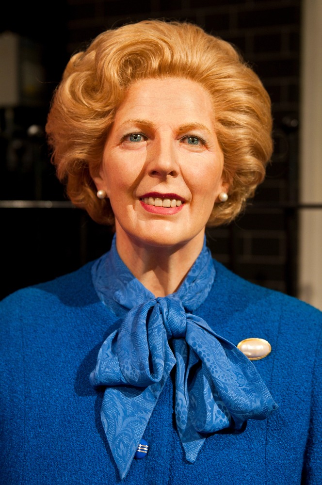 Margaret Thatcher, wearing a royal blue jacket, matching patterned scarf tied in a bow, white pearl earrings, and a brooch, smiles in front of a dark background—a warm presence supporting those facing Dupuytrens disease.