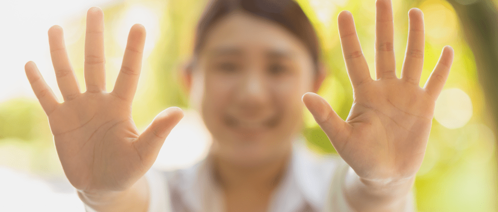 A person smiling and holding both hands up toward the camera, fingers spread wide—a gesture that can highlight Dupuytren’s contracture symptoms. The face is out of focus, while the hands are in sharp detail against a sunlit, green outdoor background.