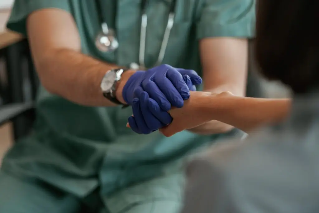 A healthcare professional wearing green scrubs and blue gloves gently holds a patient’s hand, offering comfort and support during a medical consultation about Dupuytren’s contracture symptoms.