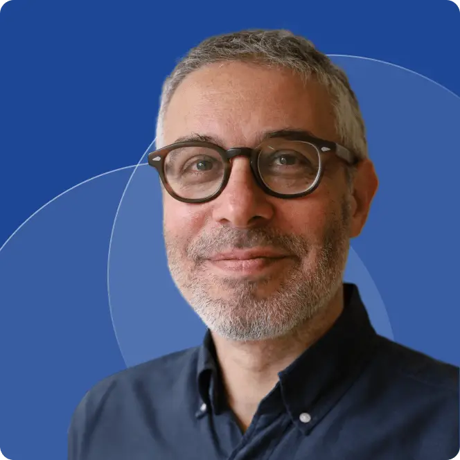A middle-aged man with short gray hair, glasses, and a trimmed beard smiles at the camera. He is wearing a dark blue button-up shirt, with a blue background featuring abstract circular shapes, symbolizing hope for Dupuytrens contracture treatment UK.