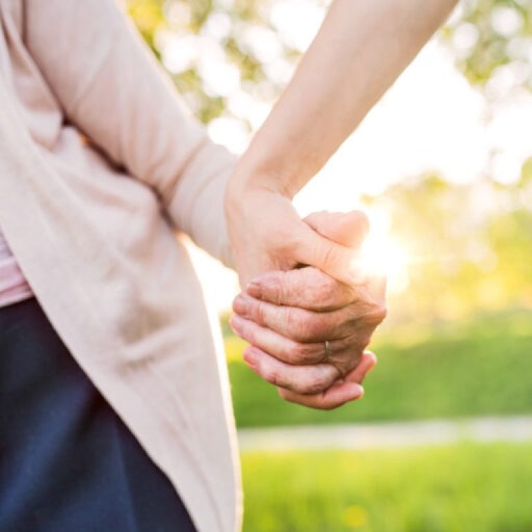 Close-up of two people holding hands outdoors with sunlight shining through, suggesting warmth and connection. The blurred green background evokes a park setting—an uplifting scene that resonates with those seeking Dupuytrens contracture treatment UK.