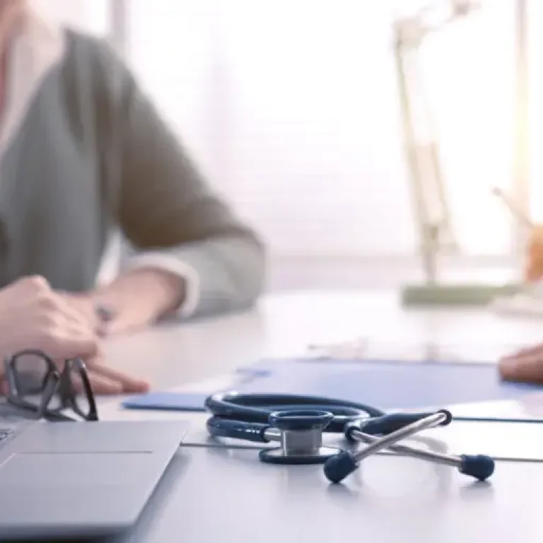 Two people sit across from each other at a desk in an office. A stethoscope, glasses, and clipboard rest on the desk, suggesting a medical consultation about Dupuytren’s contracture symptoms or causes of Dupuytren’s disease is taking place.