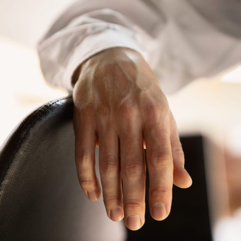 A close-up of a person’s hand, possibly affected by Dupuytren’s contracture symptoms, resting on the back of a black chair. The person is wearing a white long-sleeve shirt and the background is softly blurred.